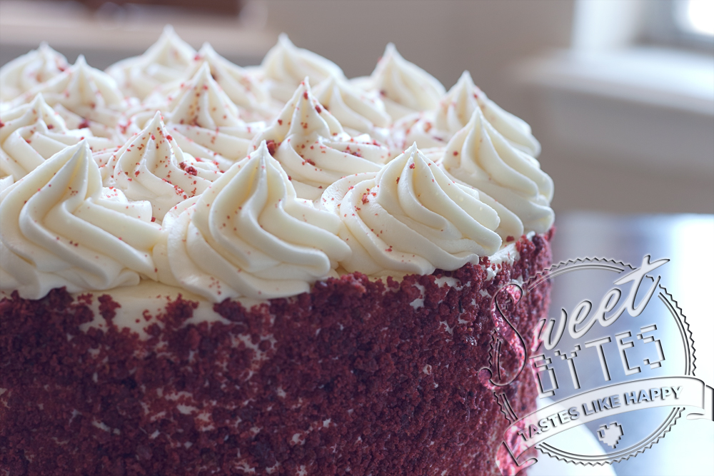 Close up picture of the top of a Red Velvet Cake with red cake crumbs covering the sides and piped cream cheese frosting swirls on top.