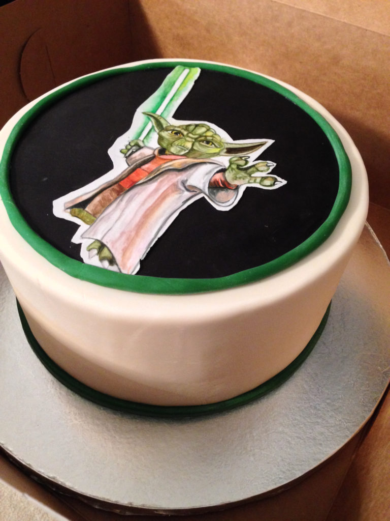 A cake covered in white fondant with hand painted yoda on top