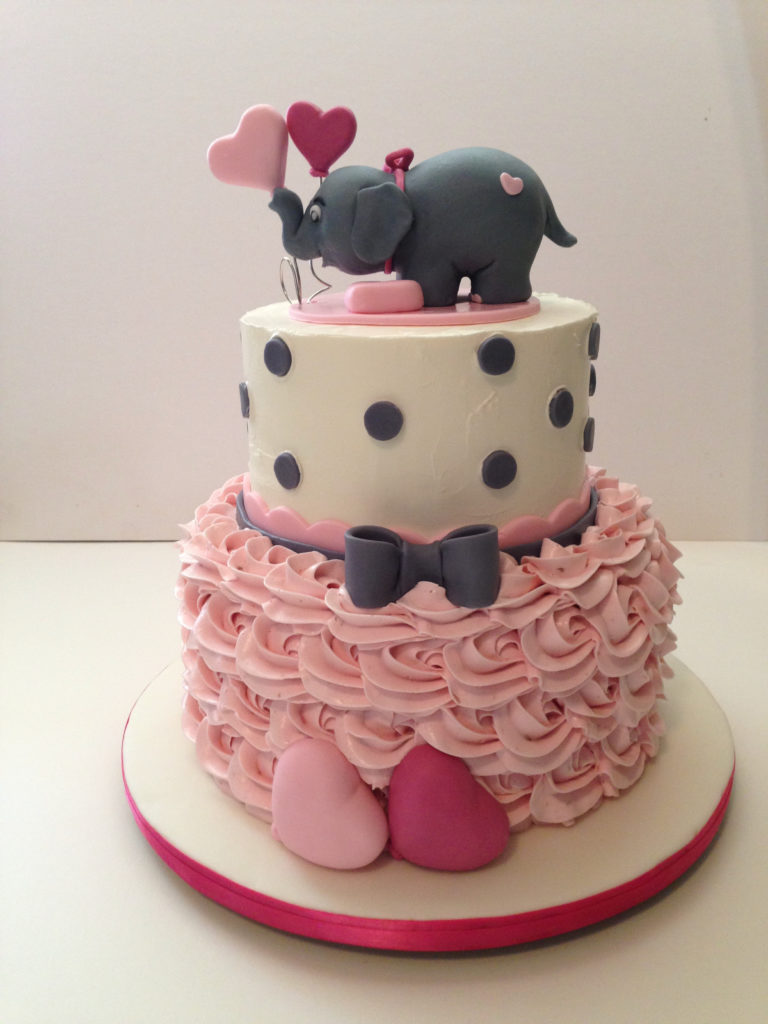 Two tiered cake with pink rosette bottom and fondant elephant holding heart balloons on top